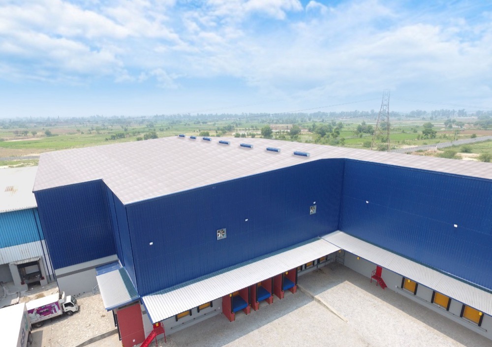 Aerial view of Connect Logistics multi-bay cold storage warehouse in Karachi — royal blue PIR roof and walls, multiple red loading docks visible at ground level, agricultural land in background